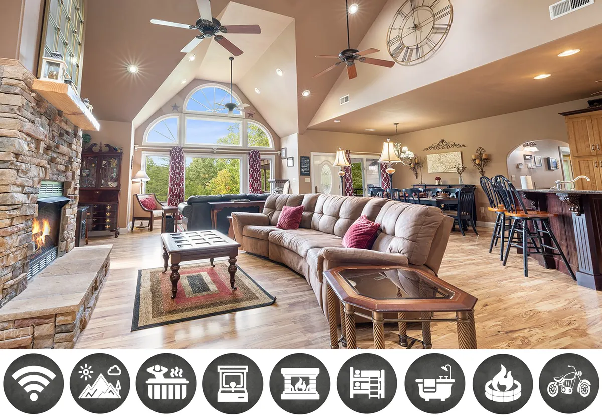 The image showcases a warm and inviting chalet interior in Sevierville, Tennessee. A grand stone fireplace serves as the centerpiece, flanked by large windows that let in natural light and offer glimpses of the Smoky Mountains. Plush, comfortable sofas are arranged for conversation or cozy relaxation, while a stylish dining area promises shared meals and laughter. The setting is ideal for family gatherings, relaxing evenings, and memorable holiday celebrations.