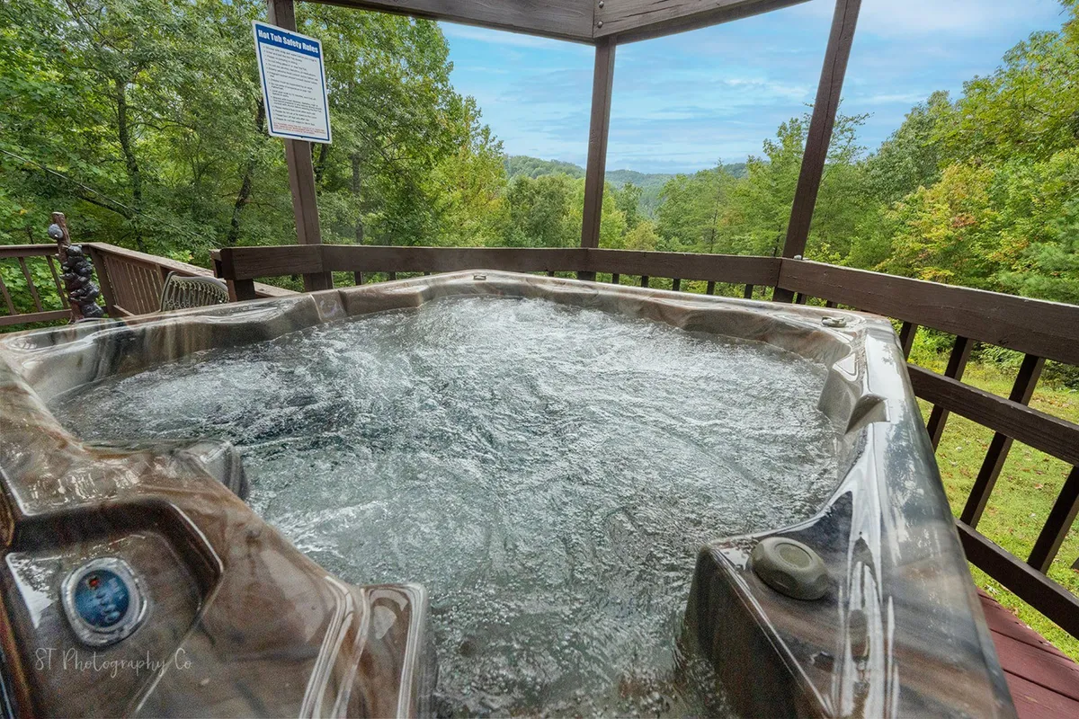 This exterior image features a hot tub situated on a wooden deck surrounded by lush greenery and offers a mountain view in the distance. The tranquil setting is ideal for couples seeking relaxation and a connection with nature.