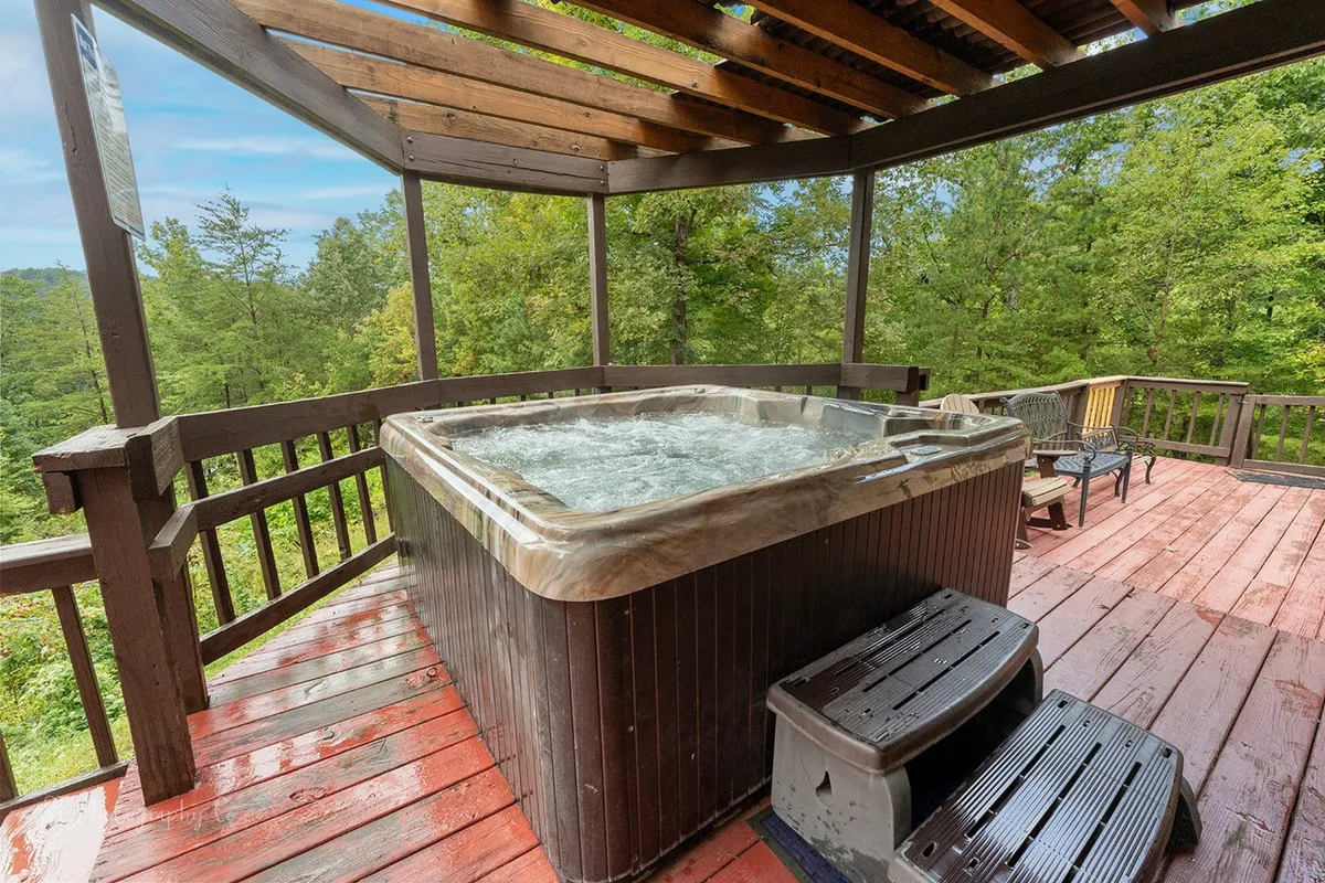 This exterior image features a hot tub situated on a wooden deck, surrounded by lush trees. The scene suggests a tranquil, communal space ideal for unwinding with friends or family. The natural setting and inviting water highlight relaxation and socialization under the open sky.