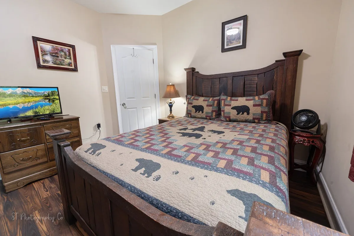 A cozy bedroom scene showcases a bed with a bear-patterned quilt, a television, a nightstand with a lamp, and a chest of drawers. The soft lighting and decor evoke a sense of warmth and privacy, ideal for unwinding after a day of adventure. The TV and plush linens are clearly visible, underlining the promise of restful comfort.