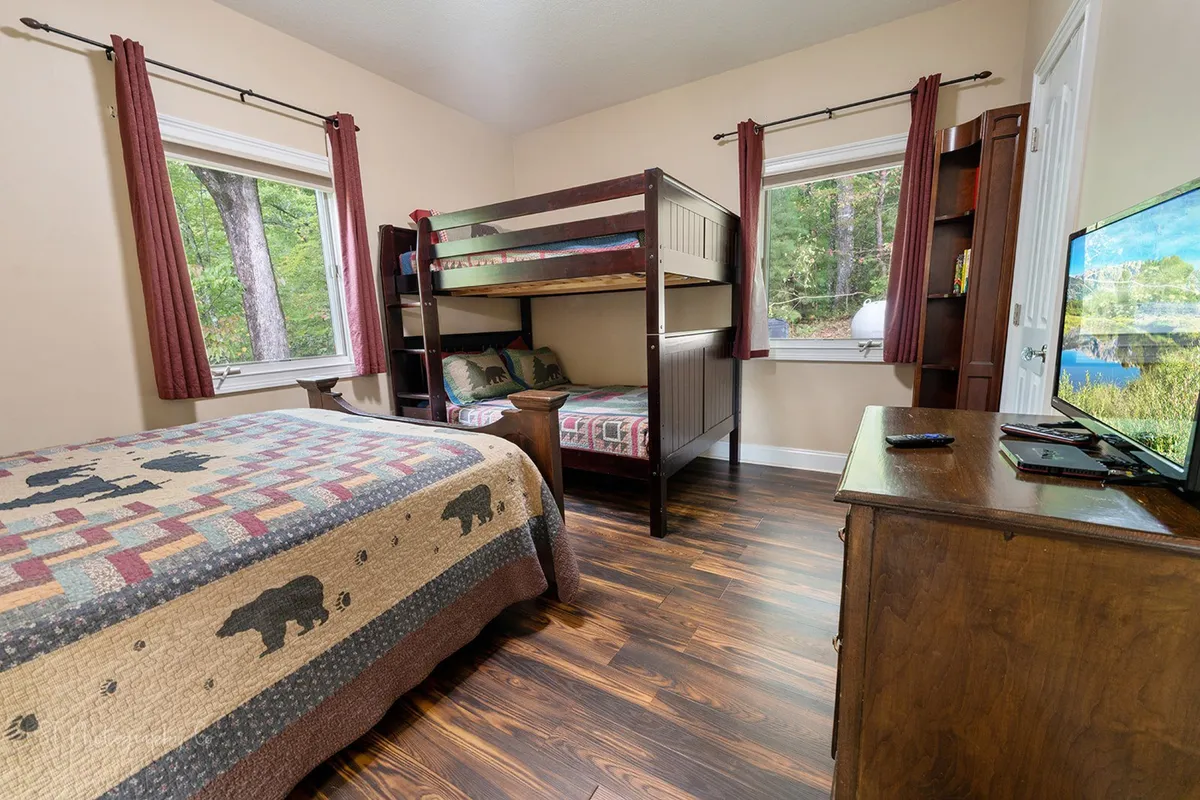 The scene shows a spacious bedroom with a queen bed and a bunk bed, complemented by a wooden dresser and a television. The layout and decor are designed for both comfort and flexibility, making it an ideal fit for families or groups traveling together. The mood is relaxed and welcoming, highlighting thoughtful amenities like the TV and ample storage.