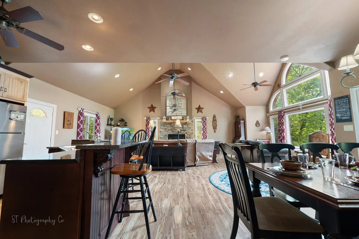 This open-concept space features a central dining table set beneath large windows, with a stone fireplace and kitchen island visible in the background. The scene exudes a sense of warmth and togetherness.