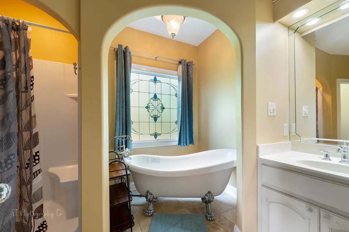 This image features a classic clawfoot bathtub set against a stained glass window, with a vanity mirror nearby. The ambiance is tranquil and elegant, suggesting a space dedicated to relaxation and rejuvenation. The bathtub is ideal for a candlelit soak or a refreshing morning ritual, perfectly matching the master suite's spa-inspired bath described in the section.